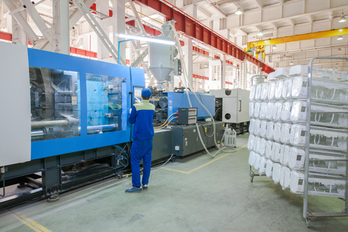 Lavoratore che utilizza una grande macchina per stampaggio a iniezione blu in una fabbrica con pile di parti in plastica pronte per la spedizione